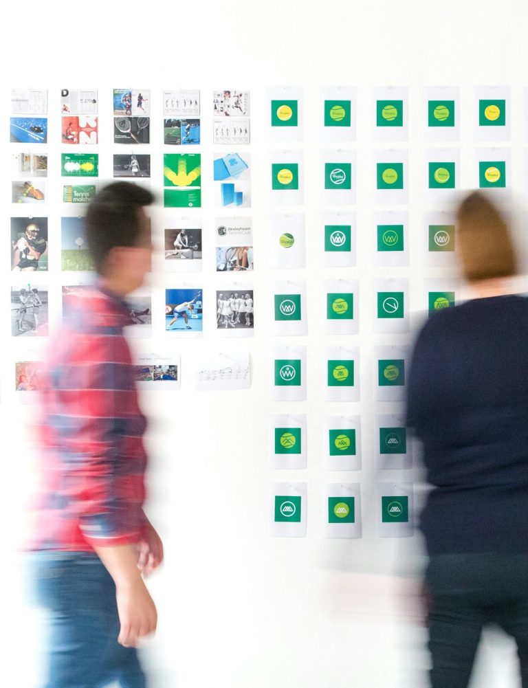 Strasser Design Ingenium – Grafikdesign, Texte, Konzepte Menschen betrachten unterschiedliche Entwürfe an einer weißen Wand.